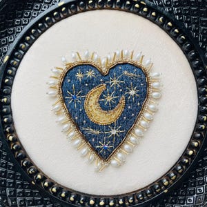 Op de afbeelding: Geborduurd hartvormig kunstwerk met een hemels thema. Het marineblauwe hart heeft een gouden halve maan en sterdetails, omringd door witte parels en gouden kralen. Het kunstwerk is ingelijst in een zwart, sierlijk frame.
