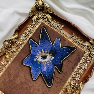 Puede incluir: Un parche bordado azul en forma de estrella con un diseño de ojo, detalles dorados y adornos de joyas. El parche está enmarcado en un marco marrón con detalles florales dorados.