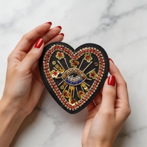 Puede incluir: Parche negro en forma de corazón con un diseño de ojo bordado. El parche presenta cuentas y lentejuelas doradas, rojas y azules. El corazón está rodeado por un borde de cuentas rojas y pequeños acentos de flores doradas.
