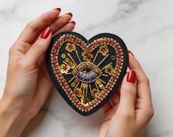 Puede incluir: Parche negro en forma de corazón con un diseño de ojo bordado. El parche presenta cuentas y lentejuelas doradas, rojas y azules. El corazón está rodeado por un borde de cuentas rojas y pequeños acentos de flores doradas.