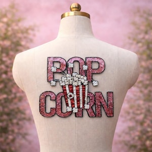 Könnte beinhalten: Ein paillettenbesetzter Aufnäher mit der Aufschrift "POP CORN" und einem Popcorn-Eimer in der Mitte. Die Buchstaben sind dunkelrosa mit silbernen Pailletten. Der Popcorn-Eimer ist rot und weiß mit silbernen Pailletten.