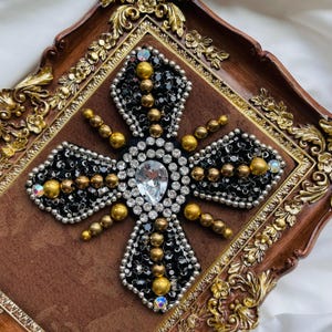 May include: A decorative cross design with a large clear crystal at the center, surrounded by black beads, gold beads, and silver accents. The cross is set on a brown velvet background within a decorative wooden frame.