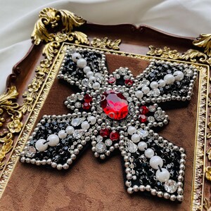 May include: A decorative cross with a large red gemstone center, surrounded by smaller red and clear crystals, black beads, and white pearls. The cross is set on a brown velvet background within a decorative gold-framed tray.
