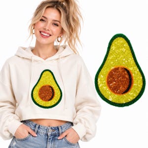 May include: A cream-colored cropped hoodie featuring a large, central avocado graphic made of yellow and green sequins. The avocado design includes a brown sequined pit with a black beaded outline, matching the beaded border of the fruit shape.