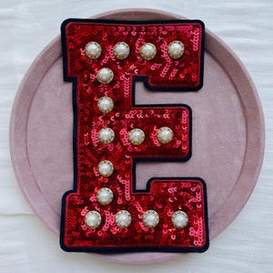 Pode incluir: Um patch da letra "E" vermelho com lantejoulas, com uma borda azul marinho e adornado com pérolas falsas. O patch é exibido em uma superfície rosa, mostrando seu design decorativo e potencial para personalização.