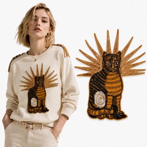 Puede incluir: Una sudadera de manga larga color crema con un gran parche bordado de un gato a rayas marrones y negras con un halo dorado de rayos de sol. Los hombros de la sudadera están decorados con flecos dorados que combinan con el diseño del gato.