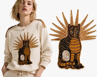 Puede incluir: Una sudadera de manga larga color crema con un gran parche bordado de un gato a rayas marrones y negras con un halo dorado de rayos de sol. Los hombros de la sudadera están decorados con flecos dorados que combinan con el diseño del gato.
