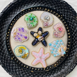 Puede incluir: Una colección de broches artesanales de conchas marinas y estrellas de mar. Los broches presentan intrincados trabajos de abalorios en varios colores, incluyendo verde, rosa, azul y blanco. Algunos están adornados con pequeñas conchas y cristales.
