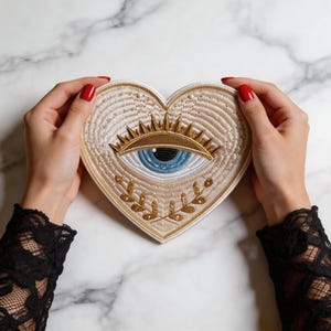 May include: A heart-shaped patch featuring intricate embroidery in shades of beige, gold, and blue. The design showcases a central eye with golden rays, surrounded by stitched patterns, small white beads, and a golden laurel branch motif.