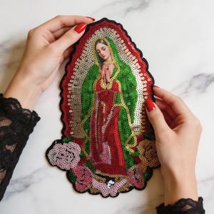 Puede incluir: Un parche bordado de forma ovalada con una representación de la Virgen de Guadalupe. El diseño está adornado con lentejuelas verdes, rojas, doradas y rosas, mostrando una figura central en oración rodeada por un borde dorado y detalles florales.