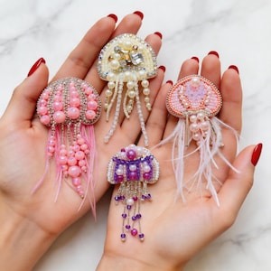 May include: Four handmade jellyfish brooches in shades of pink, white, and purple. Each brooch features beaded details, pearls, and dangling accents. Some have feather-like strands. The brooches are displayed on a marble surface.