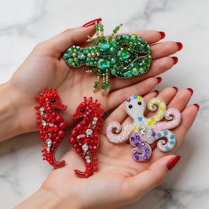 Puede incluir: Colección de broches de cuentas con criaturas marinas. Los broches incluyen una langosta verde, dos caballitos de mar rojos y un pulpo colorido. La langosta y los caballitos de mar están adornados con piedras preciosas. El pulpo tiene detalles rosas, morados, amarillos y azules.