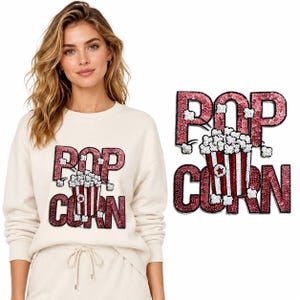 Puede incluir: Una sudadera de manga larga color crema con un gráfico de lentejuelas de un cubo de palomitas a rayas rojas y blancas con la palabra POPCORN en letras grandes de lentejuelas rosa oscuro. El diseño incluye granos de palomitas blancos.