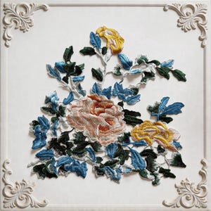 May include: Embroidered floral artwork with a large peony in pink and orange, yellow and blue flowers, and green leaves. The design is set against a white background within a white, ornate frame, creating a decorative piece for the home.