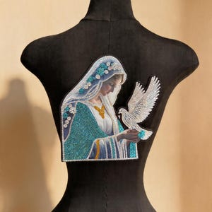 Puede incluir: Un parche decorativo que representa una figura religiosa con una túnica turquesa y blanca, sosteniendo una paloma blanca. El parche está adornado con brillantes pedrería y colocado sobre un maniquí negro.