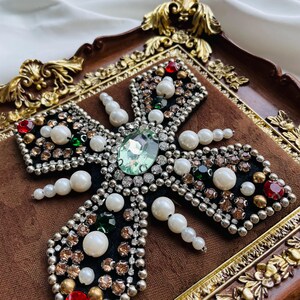 May include: A decorative cross with a black velvet base, adorned with pearls, rhinestones, and a large green gemstone. The cross is set within a brown, ornate frame, creating a luxurious and elegant aesthetic.