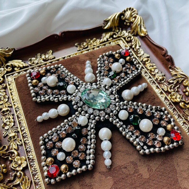 May include: A decorative, cross-shaped brooch with a central oval-cut green gemstone. The brooch is embellished with pearls, rhinestones, and red and green accents, set on a black velvet base within a brown ornate frame.