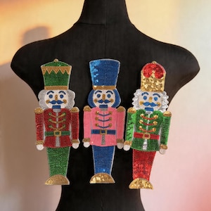 Puede incluir: Tres adornos decorativos de cascanueces. Cada adorno presenta un diseño detallado con sombrero, chaqueta y pantalón. Los cascanueces son de color rojo, verde y rosa con detalles dorados, plateados y azules. Los adornos se exhiben en un maniquí negro.