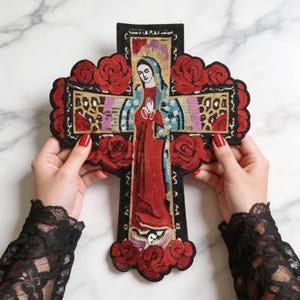 Op de afbeelding: Een grote kruisvormige geborduurde patch met een centrale figuur van de Maagd Maria in een rood en blauw gewaad. Het ontwerp is omringd door rode lovertjesrozen en gouden luipaardprint accenten op een zwarte achtergrond met een schulprand.