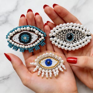 May include: Three beaded eye brooches in various designs. One has a blue and white beaded border, another features white pearls, and the third has a gold and white beaded design. The eyes are set against a neutral background.