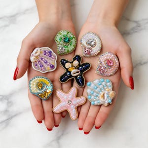 May include: A selection of beaded seashell and starfish brooches in shades of pink, blue, green, and white. Each brooch is intricately detailed with beads and small embellishments, creating a nautical theme.