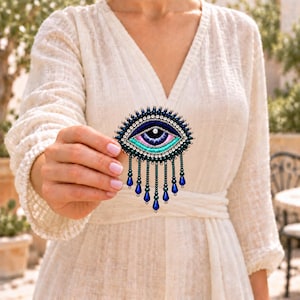May include: An eye-shaped brooch featuring dark blue, light blue, and pink embroidery with a central white bead. The piece is bordered by dark blue beads and clear crystals, with dangling dark blue teardrop beads hanging from the bottom.
