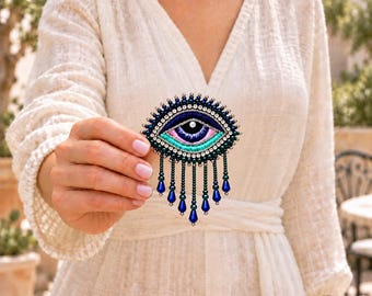 Puede incluir: Un broche con forma de ojo que presenta bordados en azul oscuro, azul claro y rosa con una cuenta blanca central. La pieza está bordeada por cuentas azul oscuro y cristales transparentes, con cuentas colgantes en forma de lágrima.