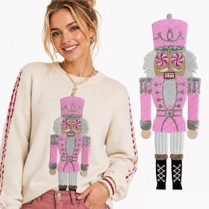 May include: A cream-colored long-sleeve sweater featuring a large, beaded nutcracker appliqué on the front. The nutcracker is decorated with pink, silver, and white sequins, peppermint candy eyes, and black boots with white cross-lacing details.