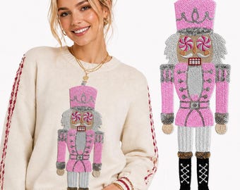 Könnte beinhalten: Ein cremefarbener Langarmpullover mit einer großen, perlenbesetzten Nussknacker-Applikation auf der Vorderseite. Der Nussknacker ist mit rosa, silbernen und weißen Pailletten, Pfefferminzbonbon-Augen und schwarzen Stiefeln mit Schnürung verziert.