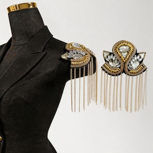 May include: Black blazer with a gold collar and decorative shoulder epaulettes. The epaulettes feature gold beading, clear crystals, and dangling gold chains. One epaulette is attached to the blazer, and the other is displayed separately.