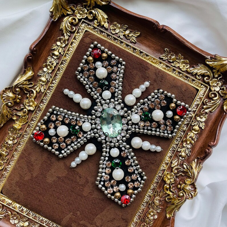 May include: A decorative cross with a central green gem, surrounded by pearls, red, and gold accents. The cross is set on a brown velvet background within a detailed, ornate, gold-trimmed frame.
