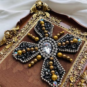 May include: A decorative, ornate cross-shaped embellishment. The design features a large, pear-shaped clear crystal at the center, surrounded by smaller clear crystals, black beads, and gold-colored beads. The embellishment is set on a brown velvet background and framed in a gold-toned, ornate border.
