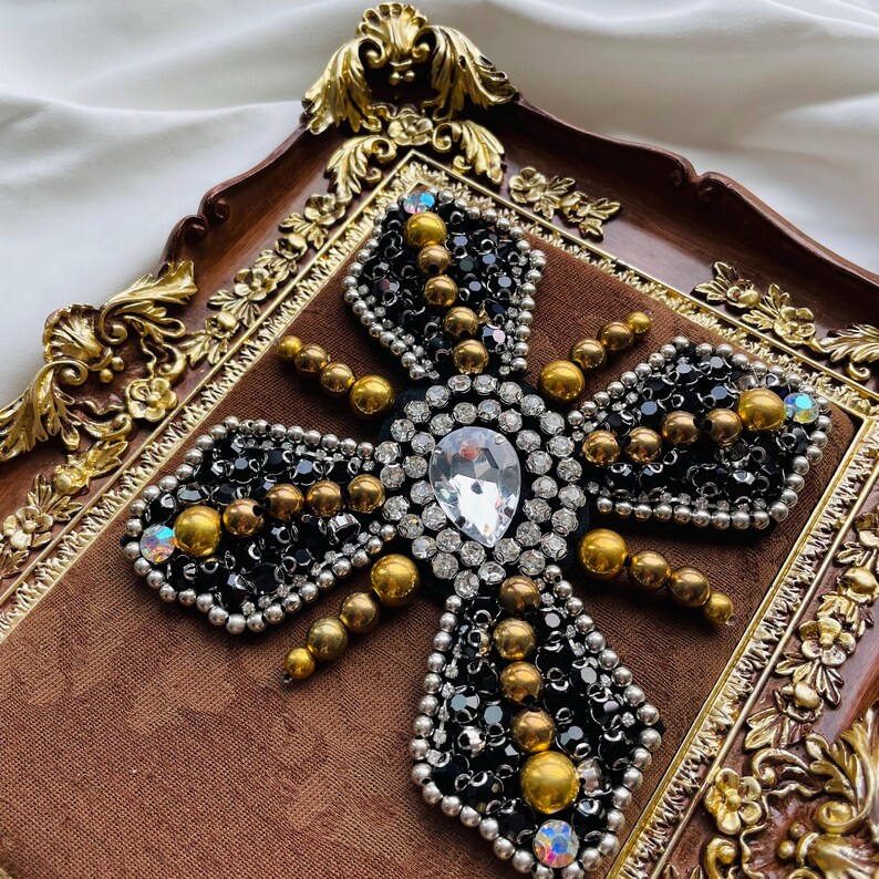 May include: A decorative, ornate cross-shaped embellishment. The design features a large, pear-shaped clear crystal at the center, surrounded by smaller clear crystals, black beads, and gold-colored beads. The embellishment is set on a brown velvet background and framed in a gold-toned, ornate border.