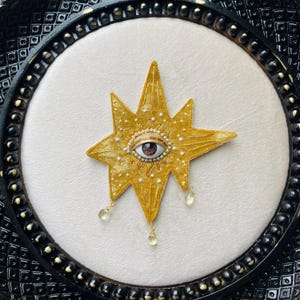 May include: A gold star-shaped decorative piece with an eye in the center, surrounded by small white pearls. The star has a textured surface and hangs from a black frame. Two teardrop-shaped gems dangle from the star.