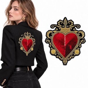 Puede incluir: Un blazer negro con un gran parche bordado en la espalda. El parche muestra un corazón rojo con una apariencia texturizada y de lentejuelas, rodeado de intrincados filigranas doradas, motivos florales y un elemento superior en forma de corona.