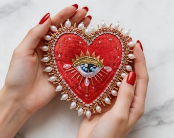 Puede incluir: Broche bordado en forma de corazón rojo con un diseño de corona y ojo dorado. El broche está adornado con perlas y cuentas doradas. El diseño está sostenido por un par de manos con esmalte de uñas rojo.
