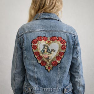 Puede incluir: Chaqueta vaquera azul claro con un parche en forma de corazón. El parche presenta una imagen religiosa de una mujer y un niño, rodeada de rosas rojas con lentejuelas y ribete dorado. La chaqueta es de estilo clásico.