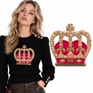 May include: A black long-sleeved ribbed sweater featuring a large gold sequined crown appliqué with a deep red velvet center. The sweater has gold buttons on the shoulder and cuffs. A separate matching crown patch is shown to the right.