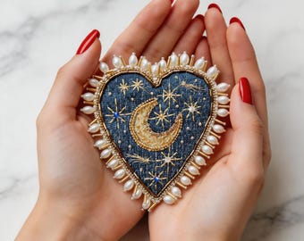 Può includere: Spilla a forma di cuore con un design celeste. La spilla presenta una base in tessuto blu scuro, adornata con stelle ricamate in oro e una luna crescente. È bordata con perle finte e perline dorate. La spilla è tenuta in due mani con smalto rosso.