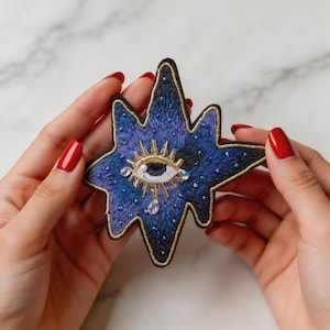 May include: Embroidered star-shaped patch in shades of blue and purple, featuring an eye design with gold accents and crystal embellishments. The patch has a black border. Held by hands with red nail polish.