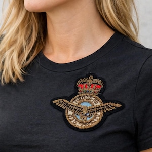 May include: Black crew neck t-shirt featuring an embroidered patch. The patch has a crown, wings, and a circular emblem with blue, gold, and red accents. The t-shirt is a solid color and appears to be made of a soft material.