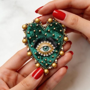 May include: A heart-shaped brooch with a green velvet base, featuring an eye design with a light green iris and black eyelashes. The eye is framed by gold and clear crystals. The brooch is edged with gold beads and small gold studs.
