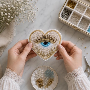Puede incluir: Parche bordado en forma de corazón con un diseño de ojo azul, detalles dorados y blancos. El parche está adornado con cuentas e hilo dorado. También se ve un plato pequeño con cuentas y un recipiente con cuentas.