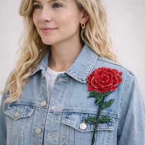 Könnte beinhalten: Hellblaue Jeansjacke mit einer großen, perlenbesetzten roten Rosenbrosche. Die Rose hat detaillierte Blütenblätter und grüne Blätter. Die Jacke hat silberne Knöpfe und ein klassisches Design.