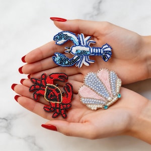 May include: A selection of beaded brooches with a marine theme. The brooches feature a blue lobster with sequins, a red crab with black detailing, and a pink and blue seashell with pearl and turquoise embellishments. The brooches are held in open hands.