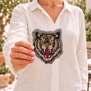 May include: A tiger head patch crafted with intricate beadwork and sequins in shades of gold, brown, black, and white. The design features a snarling expression with a prominent red mouth, all set against a sheer black mesh fabric backing.