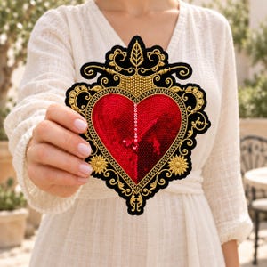May include: A heart-shaped embroidered patch featuring a central red sequined heart surrounded by intricate gold thread embroidery on a black felt base. The design includes ornate scrollwork, floral motifs, and a vertical line of white sequins.