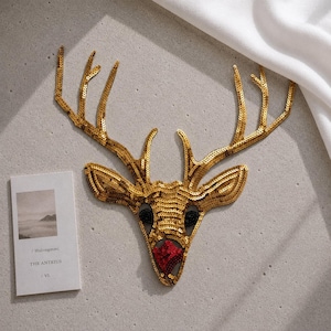 May include: A sequined deer head patch featuring gold-colored sequins covering the antlers and face, black sequined eyes, and a red sequined nose. The patch is positioned next to a rectangular white card with the text Hulongmmi THE ANTEIUS VI.