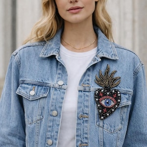 May include: Light blue denim jacket with a beaded heart-shaped patch. The patch features a blue eye, red accents, and pearl-like beads. The jacket has a classic design with button closures and pockets.
