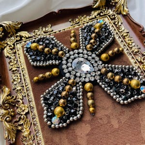 May include: A decorative butterfly design with black beads, gold accents, and a large clear gem. The butterfly is set on a brown fabric background within a decorative, ornate, brown and gold framed tray.